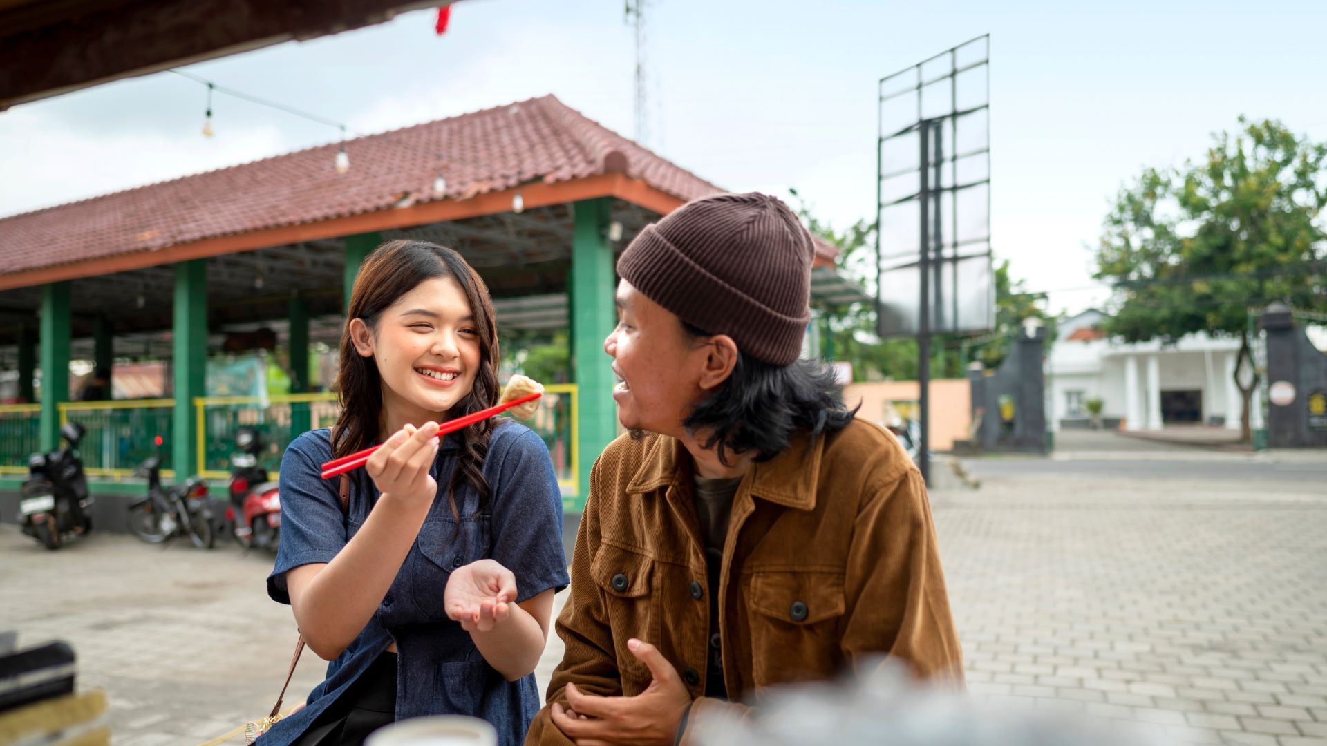 Two people at an outdoor cafe, one offers food with chopsticks, both smiling.
