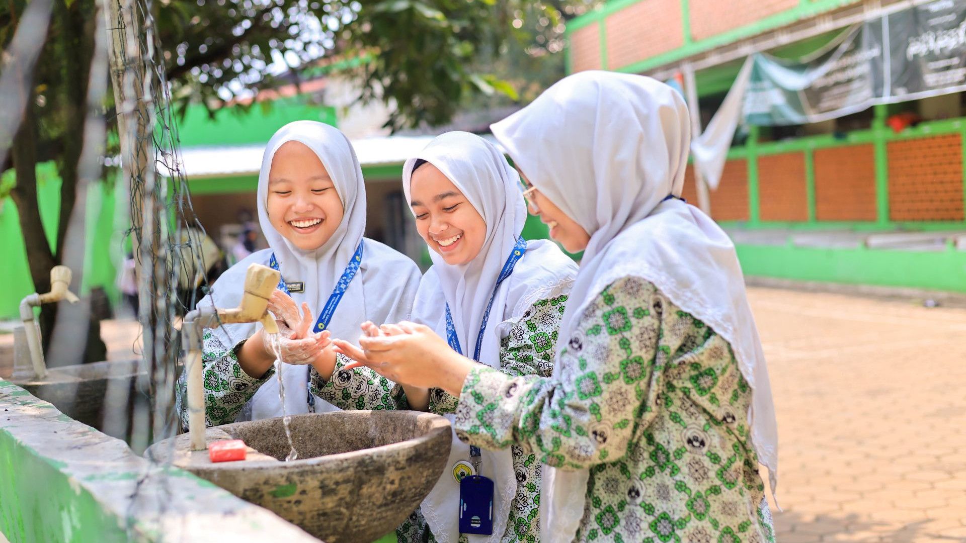 Three individuals in traditional attire are washing their hands at an outdoor sink, surrounded by a school background.