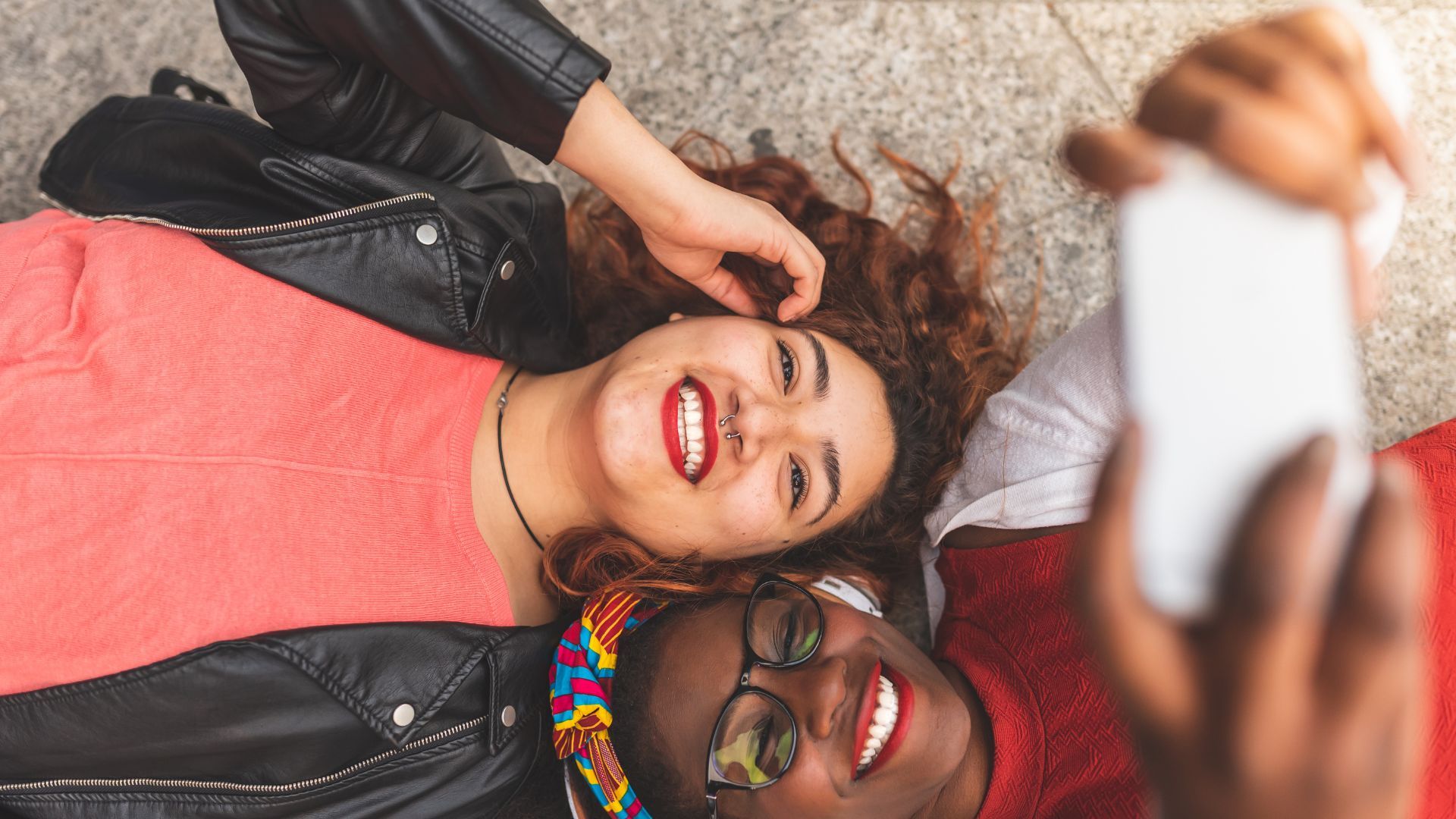 Two friends lying on the ground taking a selfie, smiling brightly.