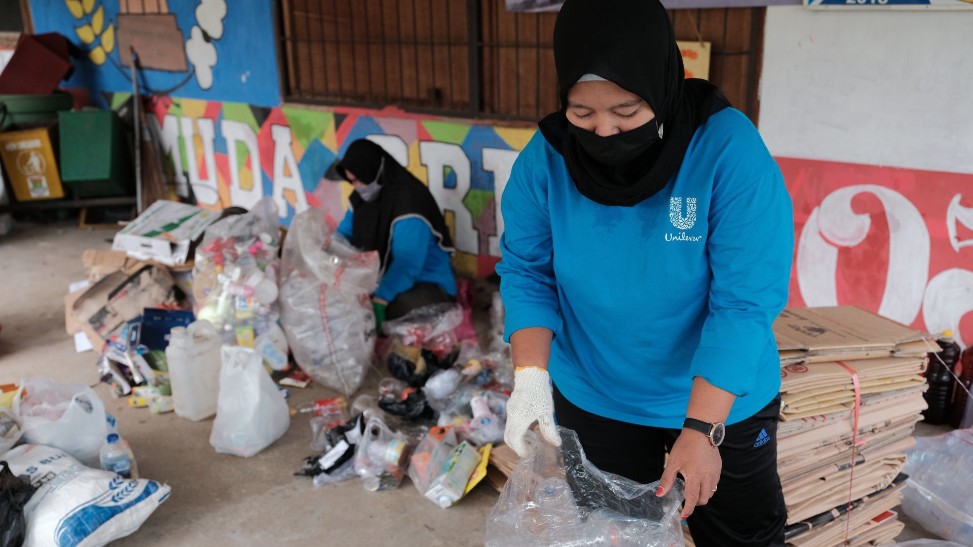 A person in a blue shirt sorts plastic waste amidst piles of recyclables in a brightly painted community space.