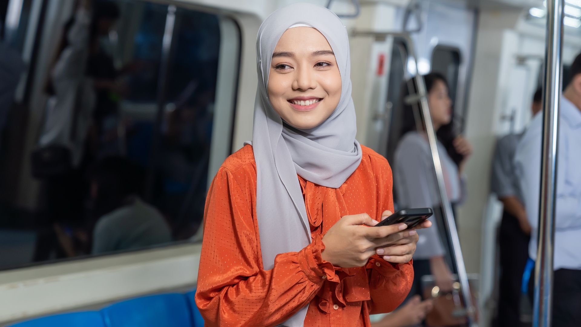 Woman in orange blouse smiling and holding phone on a train.