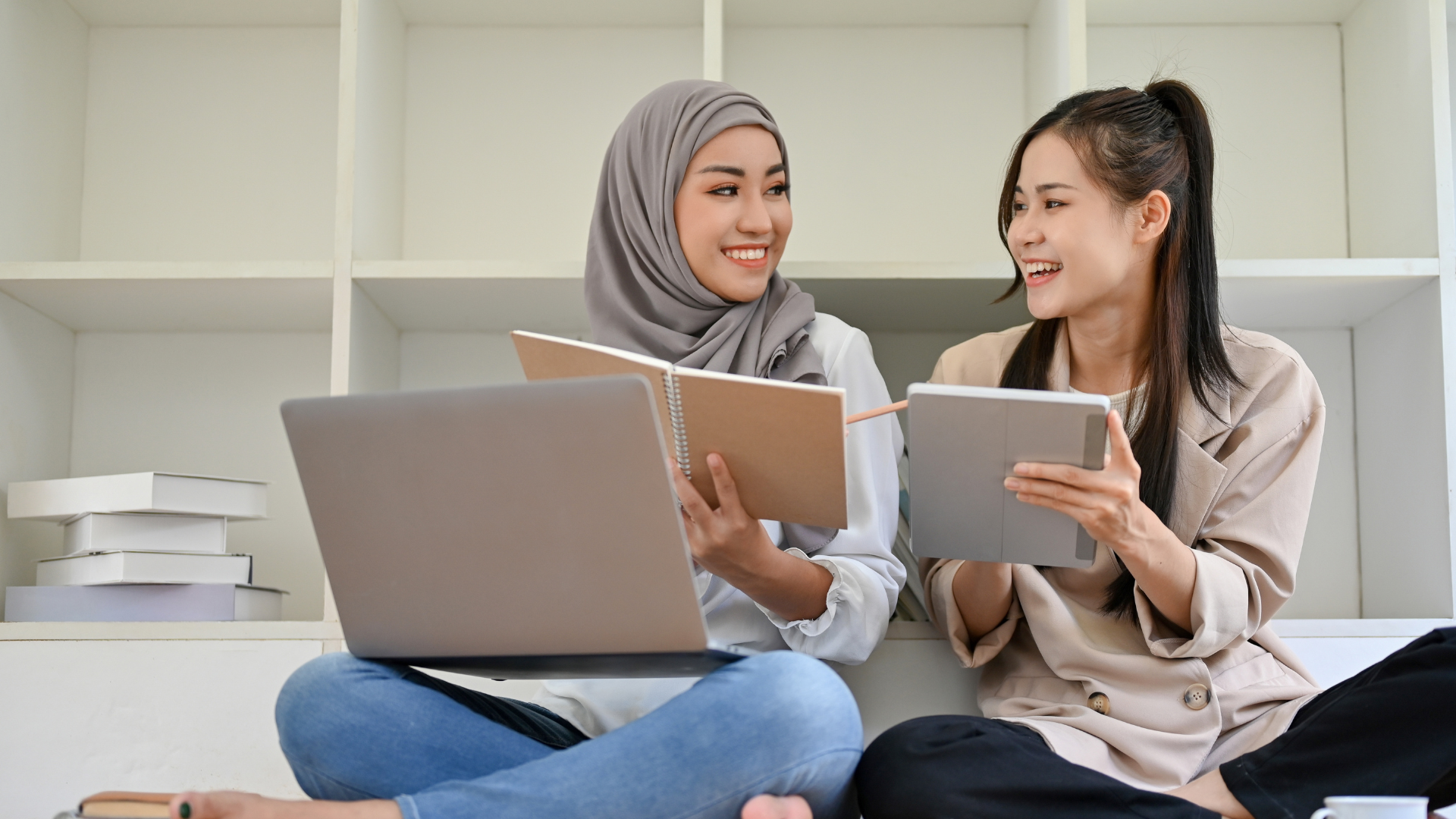 Two women smiling while studying with a laptop and notebook.