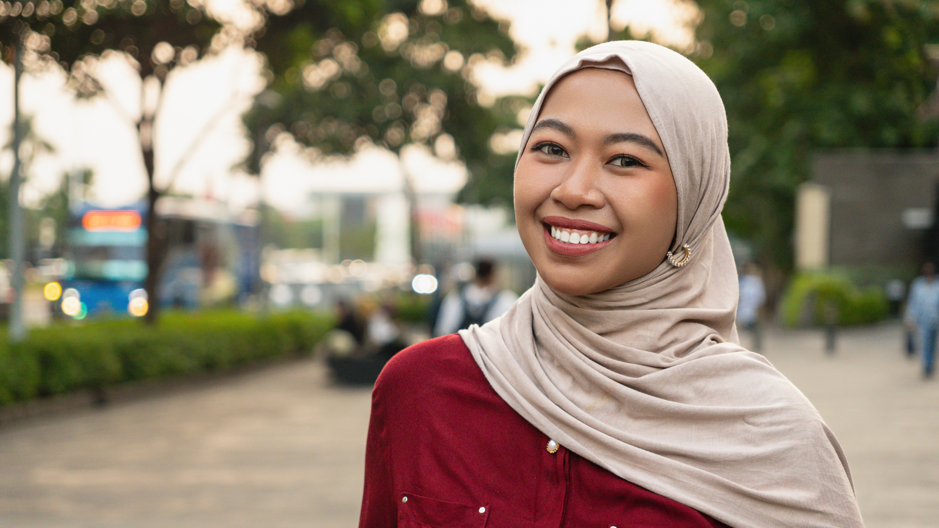 Smiling person in hijab outdoors with blurred trees and street in background.