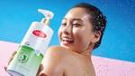 A person stands under splashing water against a bright blue background while holding a large bottle of Lifebuoy Anti Acne body wash in the foreground. The bottle has white and green packaging and features aloe vera imagery.