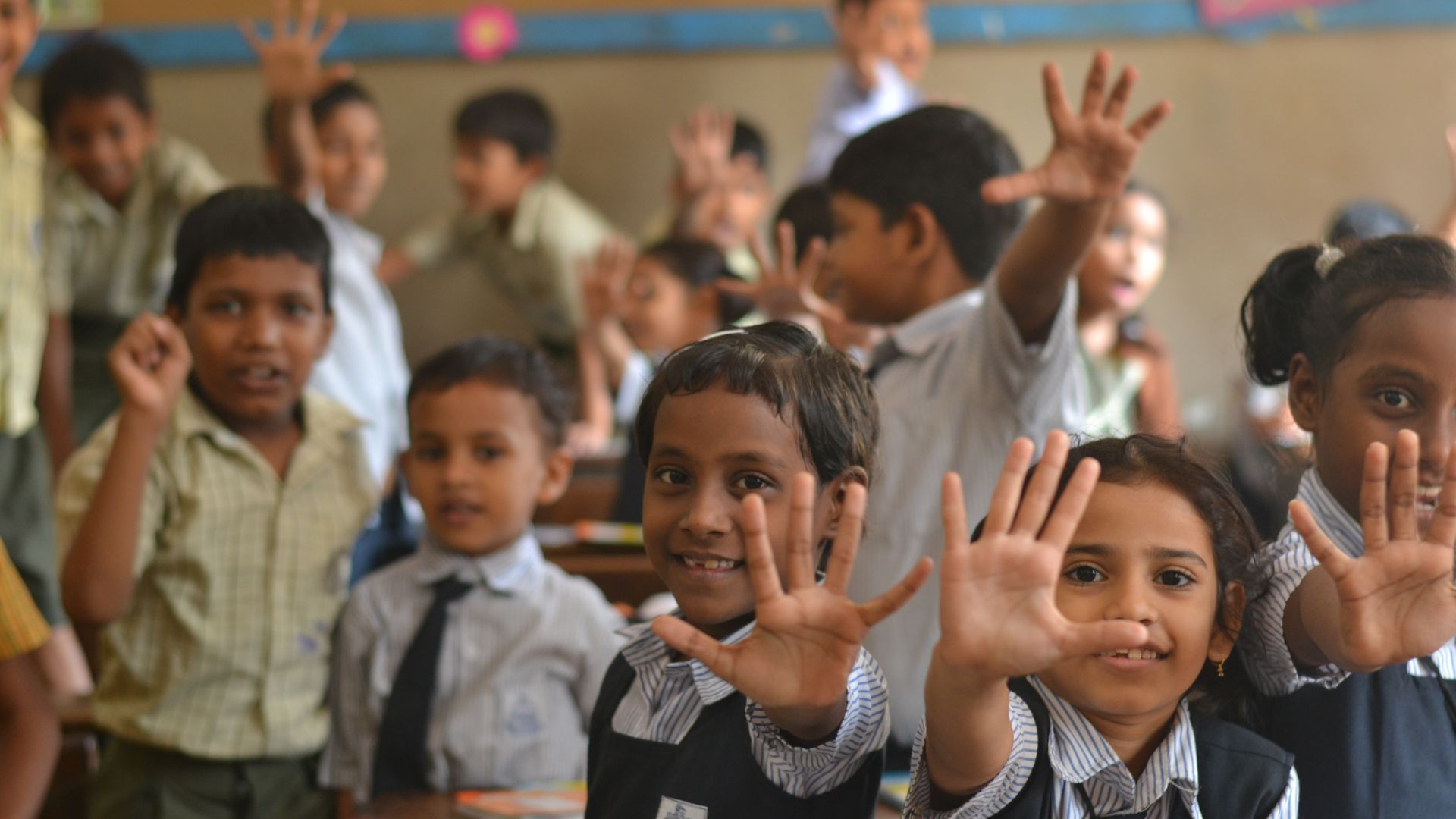 Children in a classroom raise their hands and smile at the camera. They have taken part in a Lifebuoy handwashing lesson, learning how to wash their hands with soap.