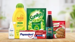 Assorted household and personal care products, including bottles and packets of brands like Zwitsal, Sunlight, Dove, Pepsodent, Bango, and Royco, arranged on a wooden surface with a blurred kitchen background.