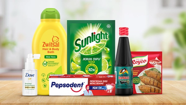 Assorted household and personal care products, including bottles and packets of brands like Zwitsal, Sunlight, Dove, Pepsodent, Bango, and Royco, arranged on a wooden surface with a blurred kitchen background.