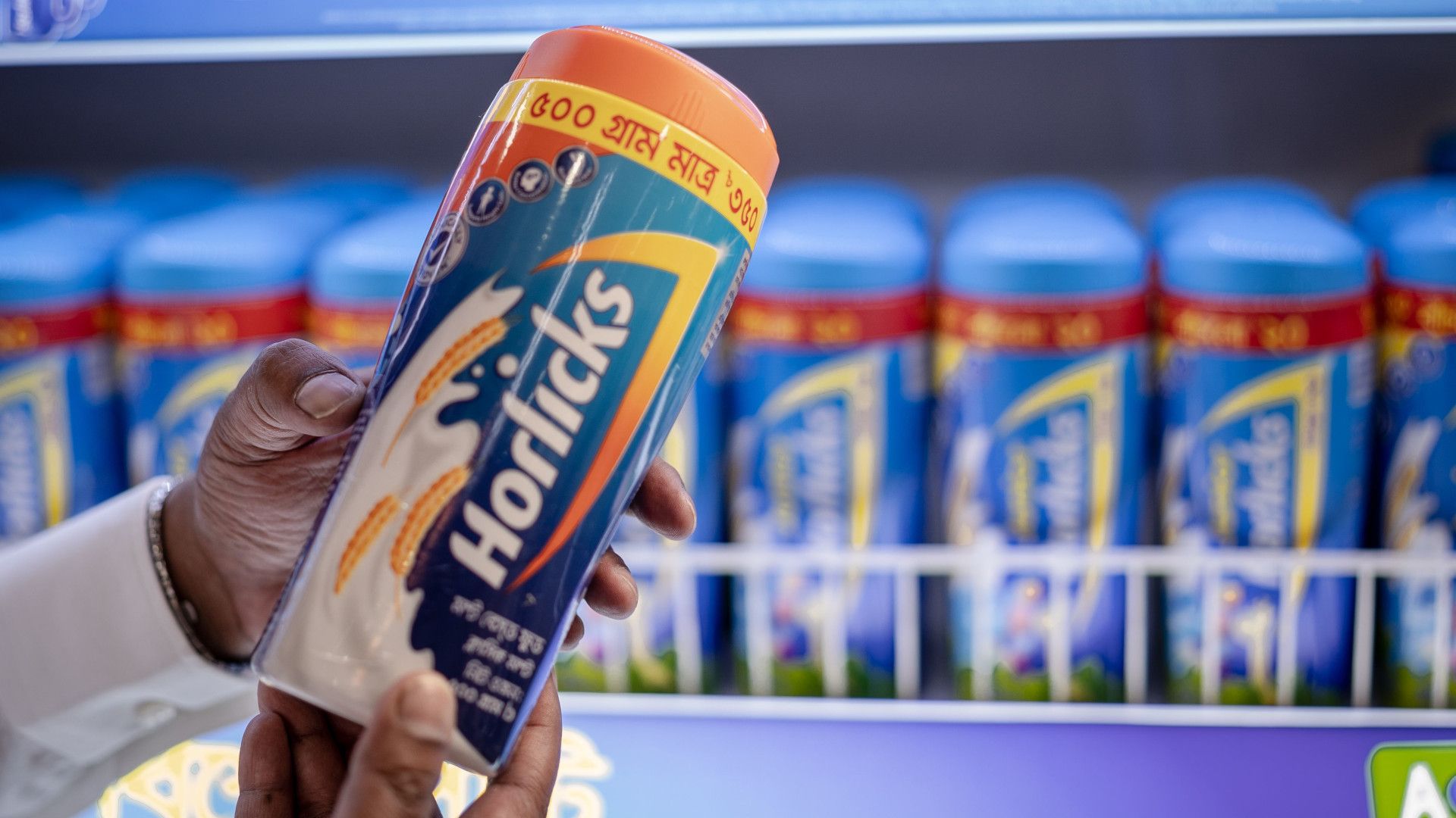 Someone holding a 500g Classic Horlicks and a shelf full of Horlicks in the background