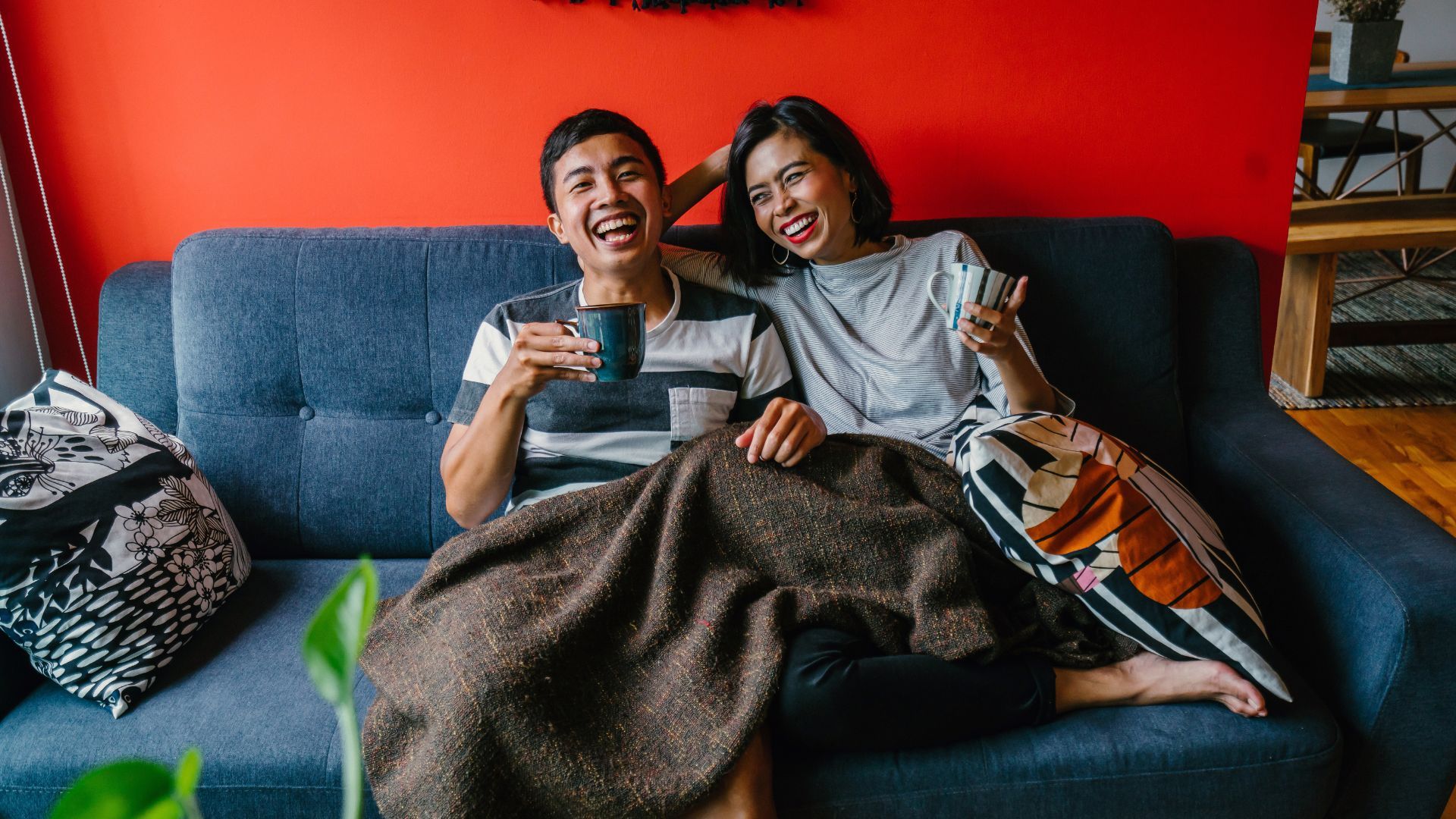 Couple sitting on a sofa, laughing and holding mugs under a blanket.
