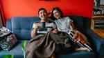 Couple sitting on a sofa, laughing and holding mugs under a blanket.